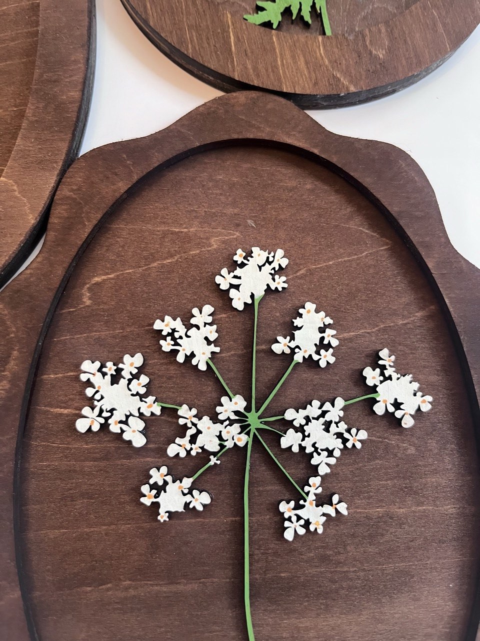 Close view of white wooden flowers and green stems against a dark wood panel, showing the grain