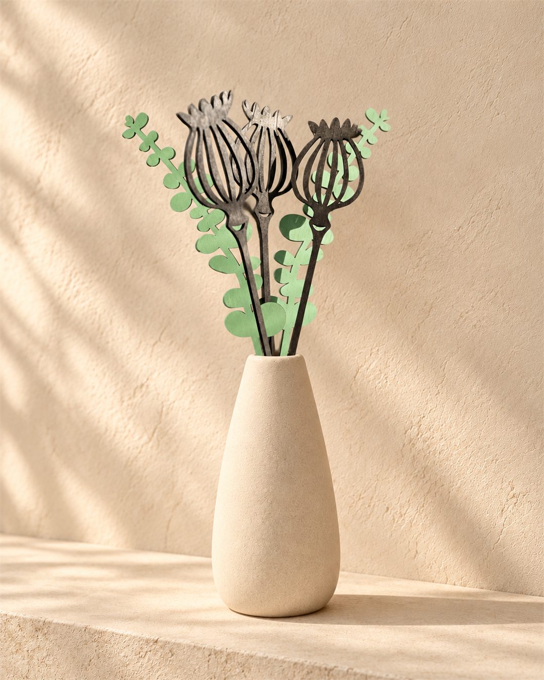 Three dark wooden seedpod forms with soft green leaves arranged in a pale vase against a warm plaster wall